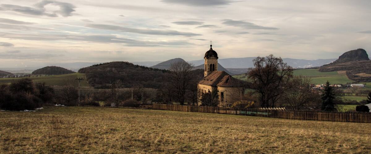 A church with a bell tower, surrounded by rolling hills and a cloudy sky.