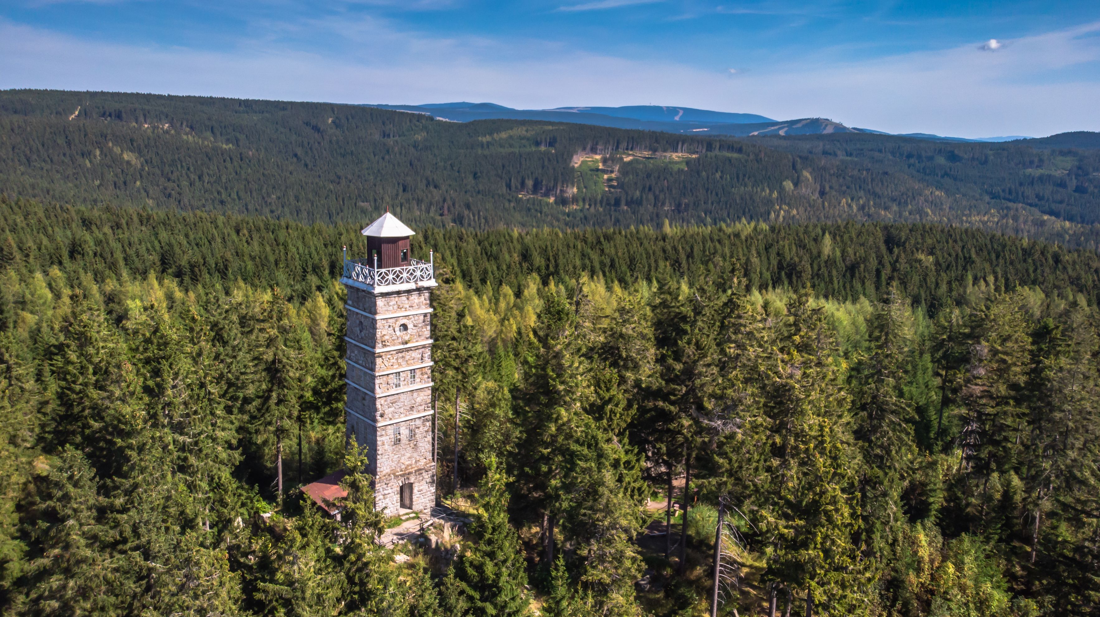 Una torre de piedra con techo cónico en medio de un bosque frondoso, rodeada de colinas ondulantes y un cielo despejado.