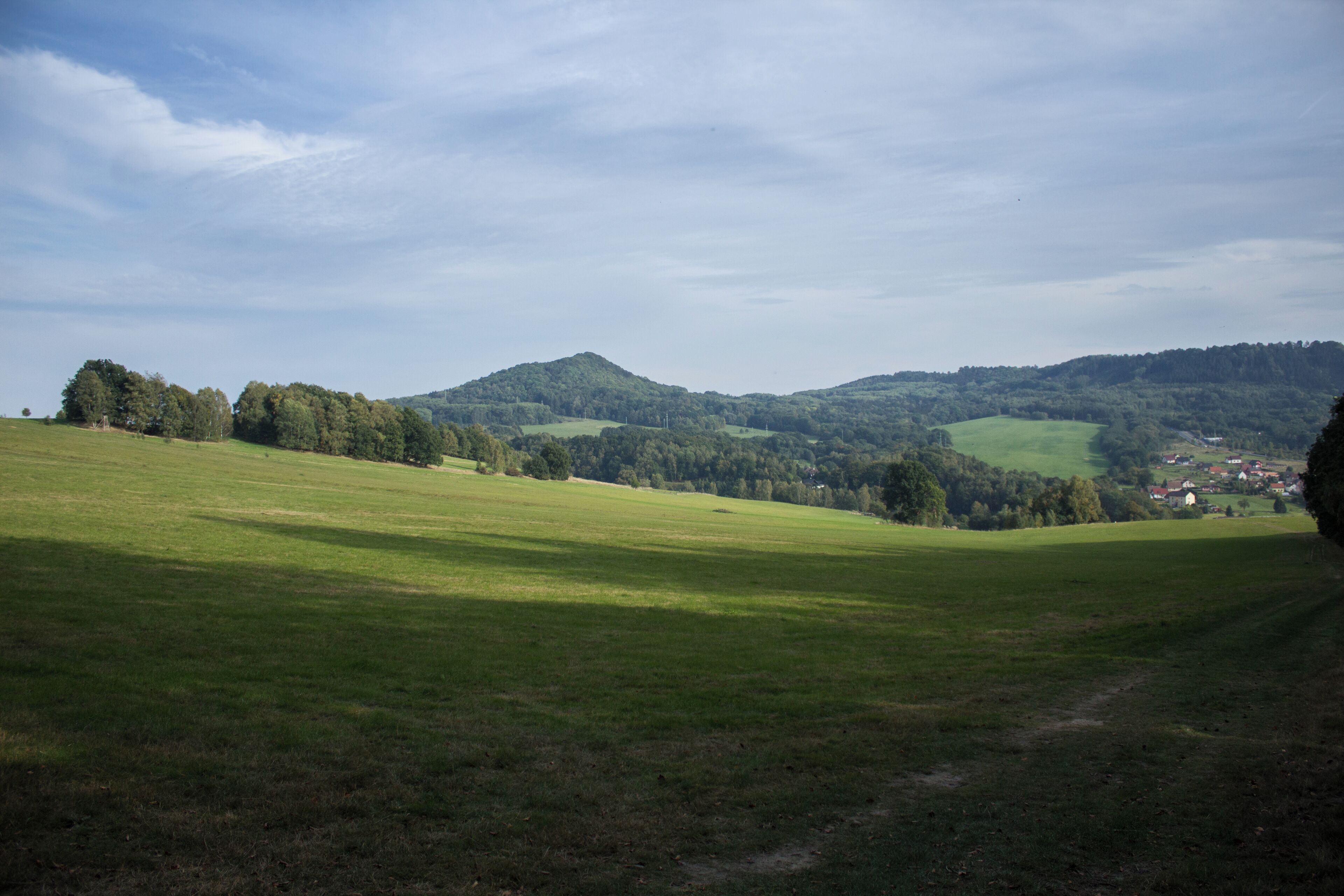 A green field with a path leading to a village in the distance, surrounded by hills and trees.