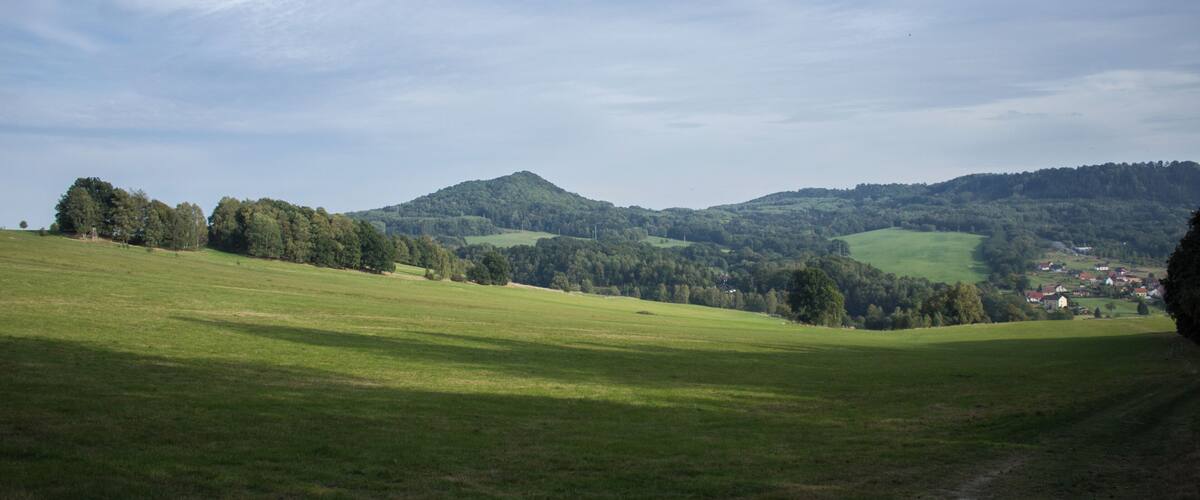 A green field with a path leading to a village in the distance, surrounded by hills and trees.