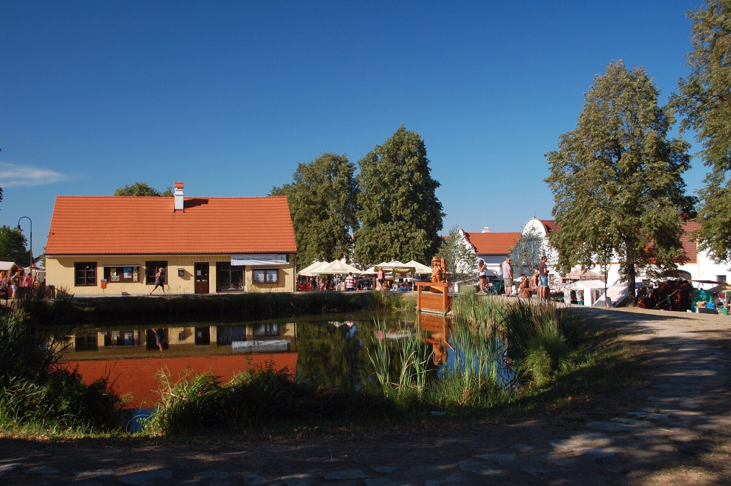 A riverside town with a red-roofed building, a pond reflecting the scenery, and people gathered around outdoor seating areas.