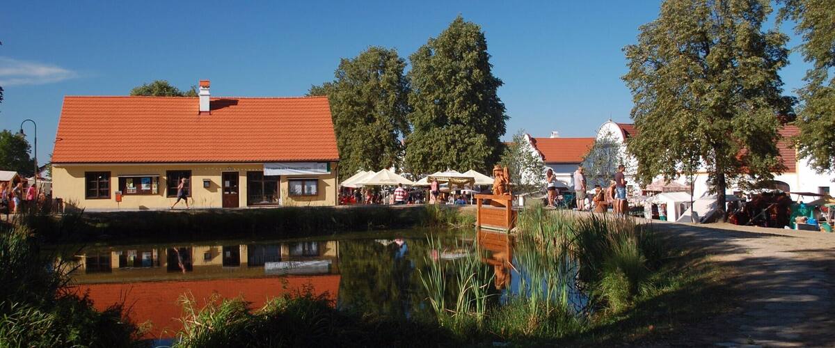 A riverside town with a red-roofed building, a pond reflecting the scenery, and people gathered around outdoor seating areas.