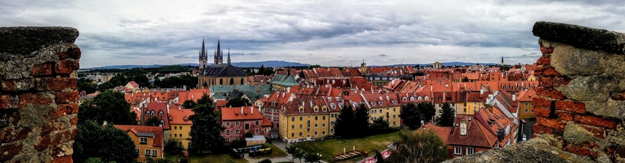 Uma cidade histórica com edifícios de telhados vermelhos, uma igreja imponente com duas torres e um cenário montanhoso ao fundo.
