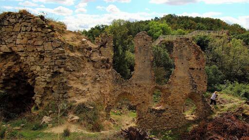 Ruinas de piedra antiguas con un puente, rodeadas de vegetación y árboles.