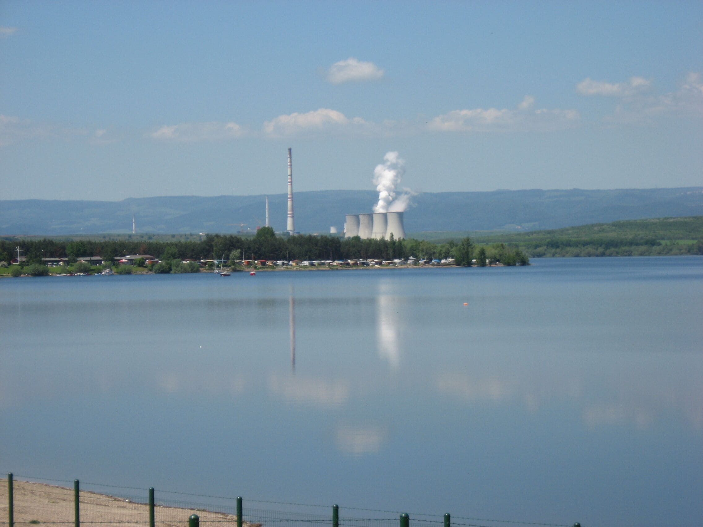 A nuclear power plant by a calm lake with smoke rising from its cooling towers.