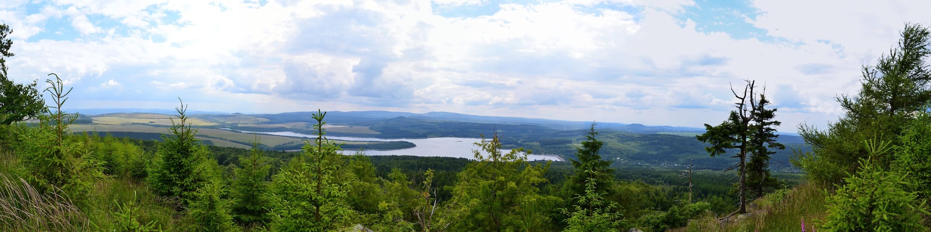 Panoramablick vom Gipfelrand des Jelení hora von Süden nach Nordwest. Im Hintergrund ist das Fichtelberg-Keilberg-Massiv, davor der Velký Špičák und im Vordergrund Stausee sowie Damm der Talsperre Preßnitz erkennbar. Unterhalb des Dammes liegt die Ortschaft Kryštofovy Hamry. Im Süden lässt sich zudem der Mědník sowie der Steilabbruch des Gebirges zum Egergraben hin erkennen.