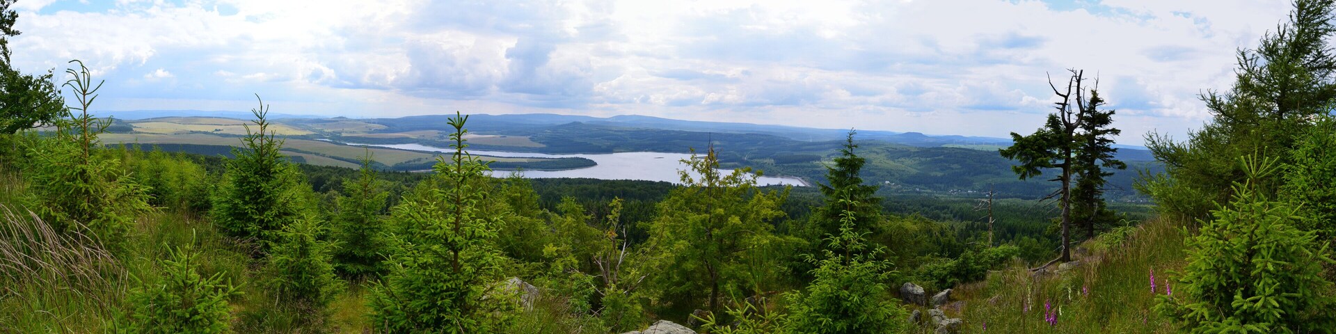 Panoramablick vom Gipfelrand des Jelení hora von Süden nach Nordwest. Im Hintergrund ist das Fichtelberg-Keilberg-Massiv, davor der Velký Špičák und im Vordergrund Stausee sowie Damm der Talsperre Preßnitz erkennbar. Unterhalb des Dammes liegt die Ortschaft Kryštofovy Hamry. Im Süden lässt sich zudem der Mědník sowie der Steilabbruch des Gebirges zum Egergraben hin erkennen.