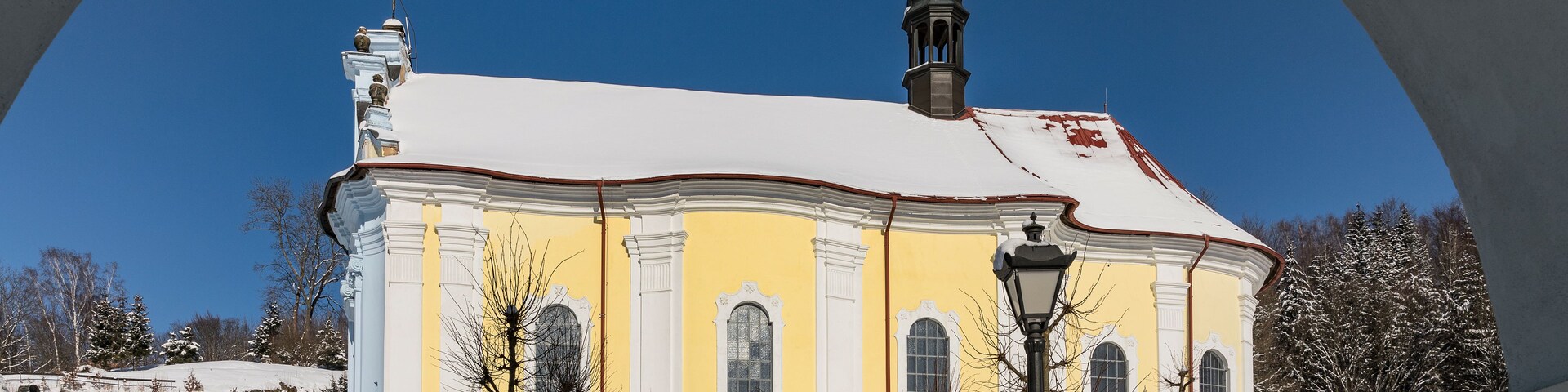 Eine schneebedeckte Kirche mit gelber Fassade und einem Glockenturm, eingefasst von einem Bogen.
