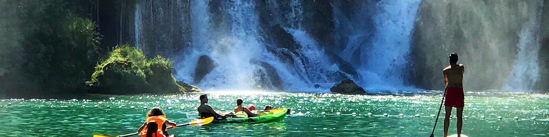 Uma cascata, caiaqueiros e pessoas em pranchas de remo em águas cristalinas.