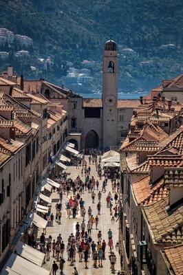 A bustling square with a prominent clock tower, surrounded by historic buildings with terracotta roofs.