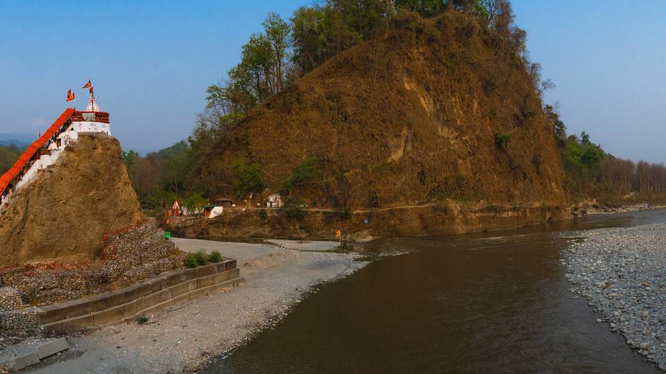 A riverbank with a large rock formation, a small shrine, and a bridge.