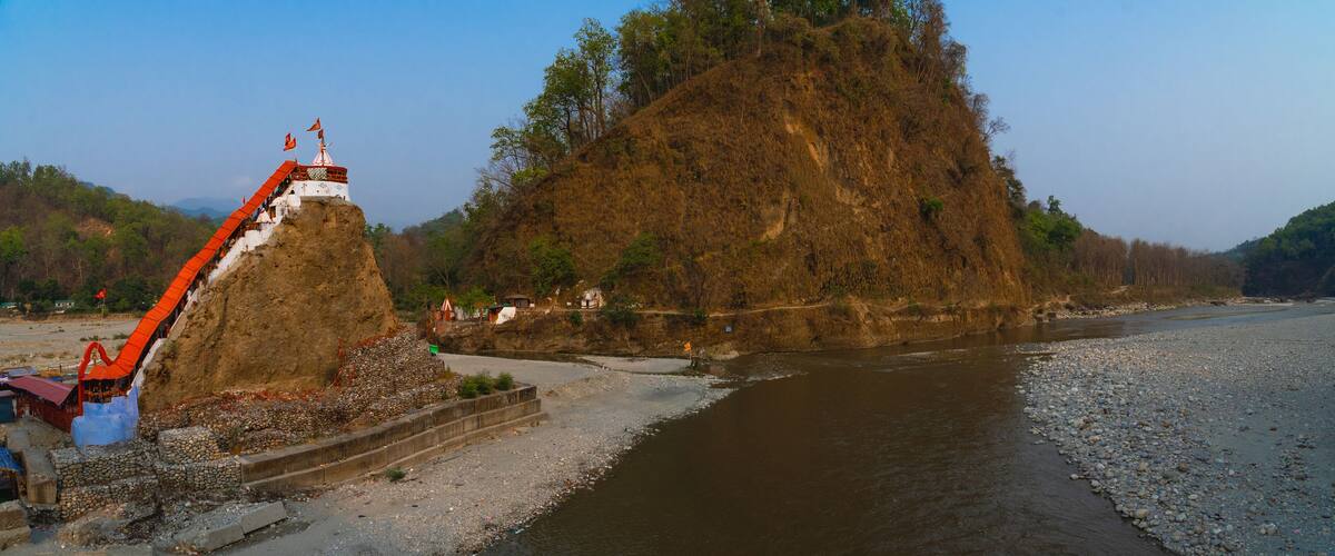 A riverbank with a large rock formation, a small shrine, and a bridge.
