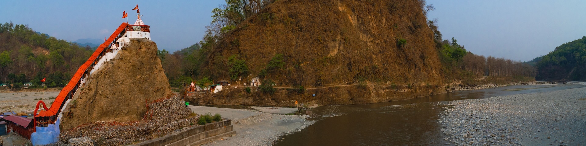 A riverbank with a large rock formation, a small shrine, and a bridge.