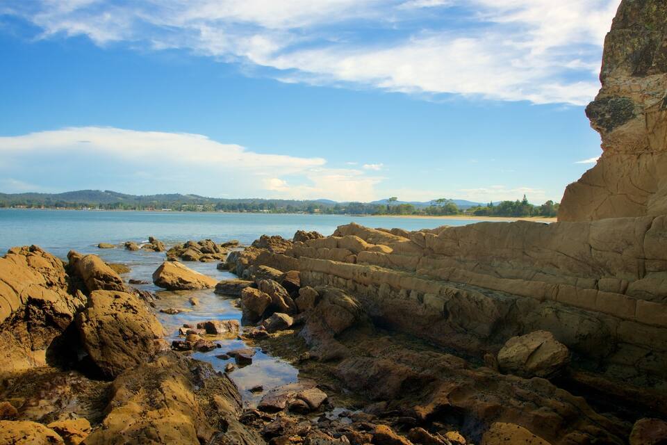 A rocky shoreline with a cliff, calm water, and distant hills.