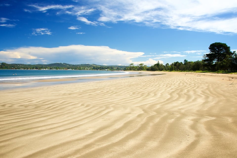 A sandy beach with gentle waves, a clear blue sky, and a forested hillside in the background.