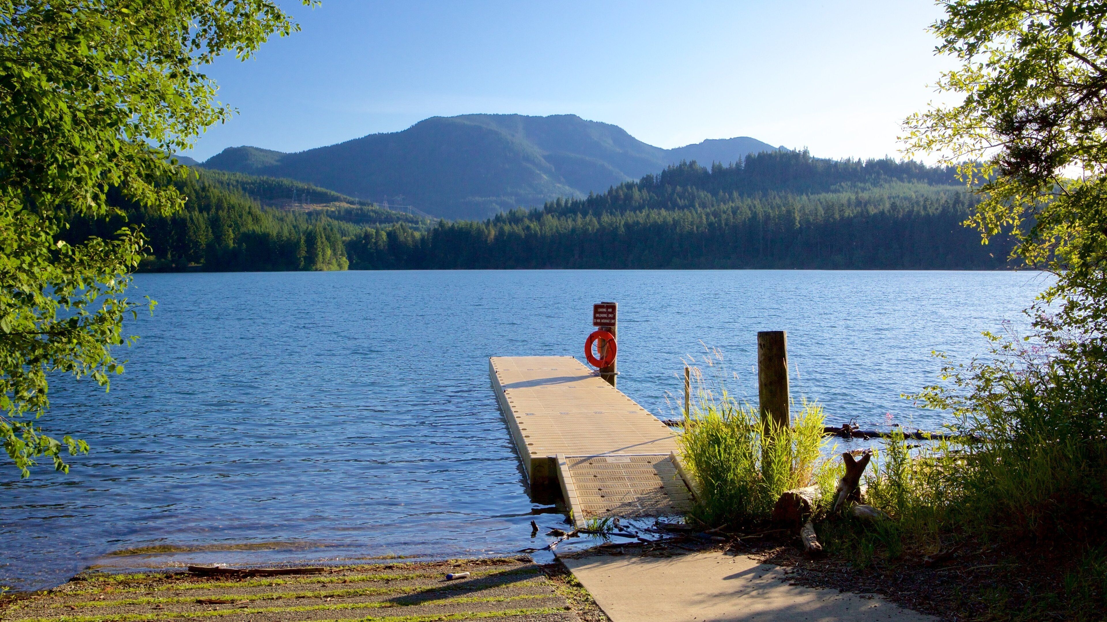 A wooden dock extending into a calm lake, surrounded by lush greenery and mountains in the background.