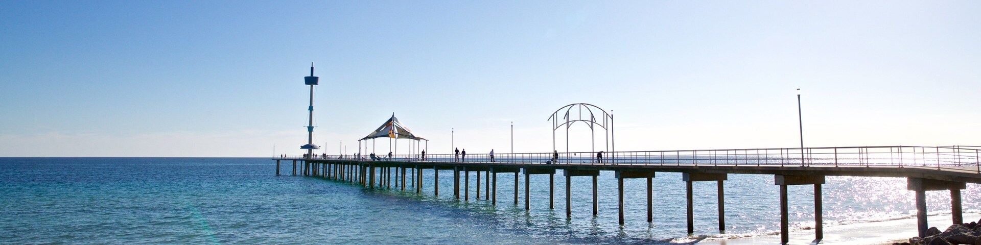 Una playa con un muelle que se adentra en el océano, un faro y un cielo azul despejado.