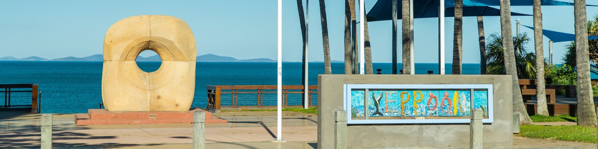 Escena costera con una gran escultura de concreto, palmeras y banderas, incluyendo la bandera australiana.