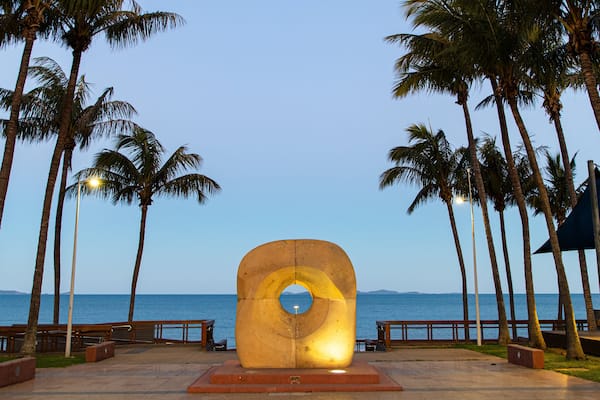 Une sculpture en forme de grand cercle jaune, avec un cercle plus petit au centre, entourée de palmiers et de bancs, et offrant une vue sur l’océan.