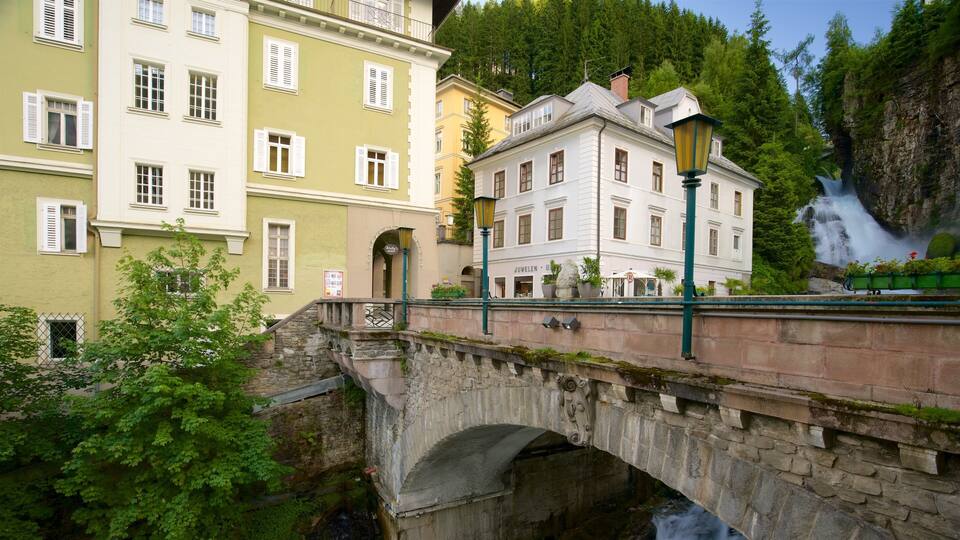 A stone bridge over a stream, surrounded by buildings with white and yellow facades, and a waterfall in the background.