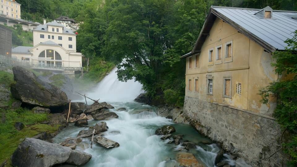A waterfall flowing through a rocky stream, surrounded by lush greenery and a historic building with a yellow facade.