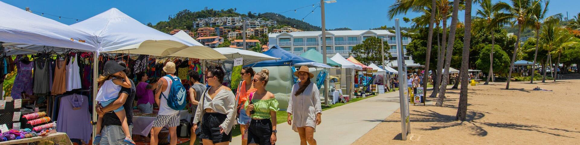 A sunny beachside market with people walking and stalls selling clothes.