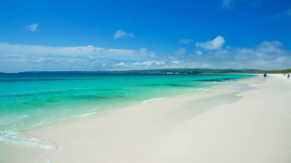 A beach with clear turquoise waters, white sand, and a few people walking along the shore.