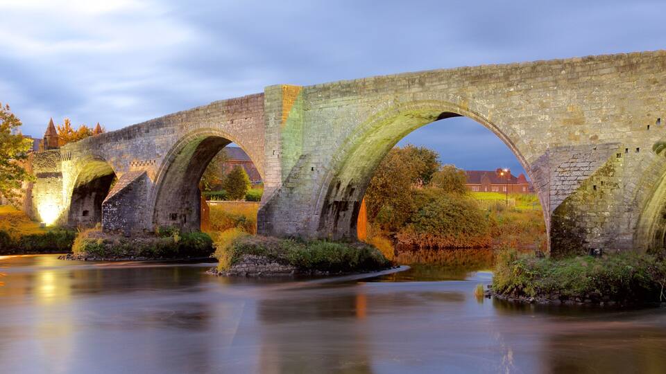 A stone bridge with multiple arches over a calm river, surrounded by greenery and historic buildings.