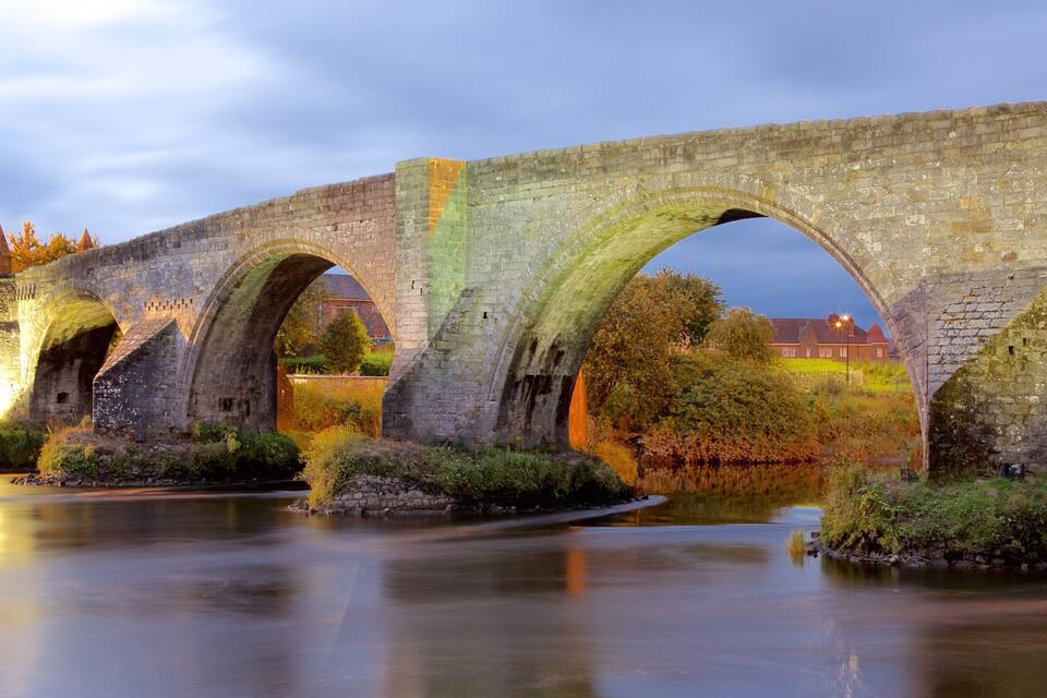 A stone bridge with multiple arches over a calm river, surrounded by greenery and historic buildings.