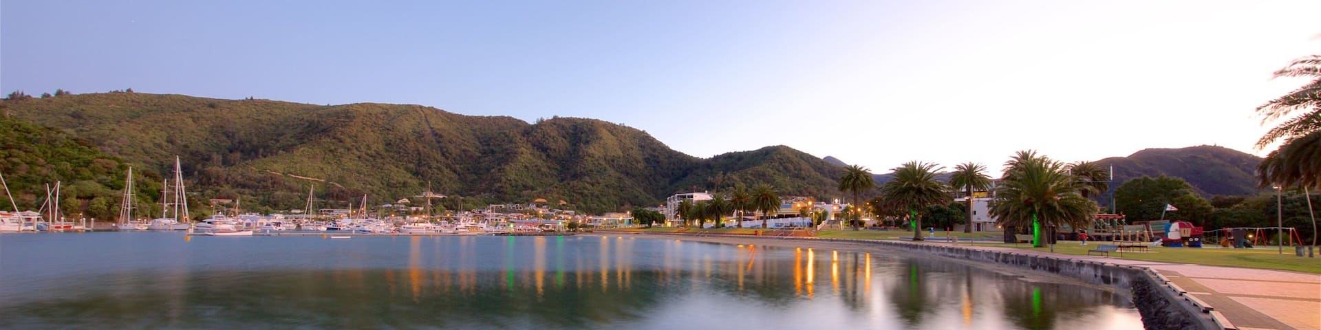 A calm lake with a wooden bench in the foreground, surrounded by a paved walkway. In the background, there are hills and a marina with boats.