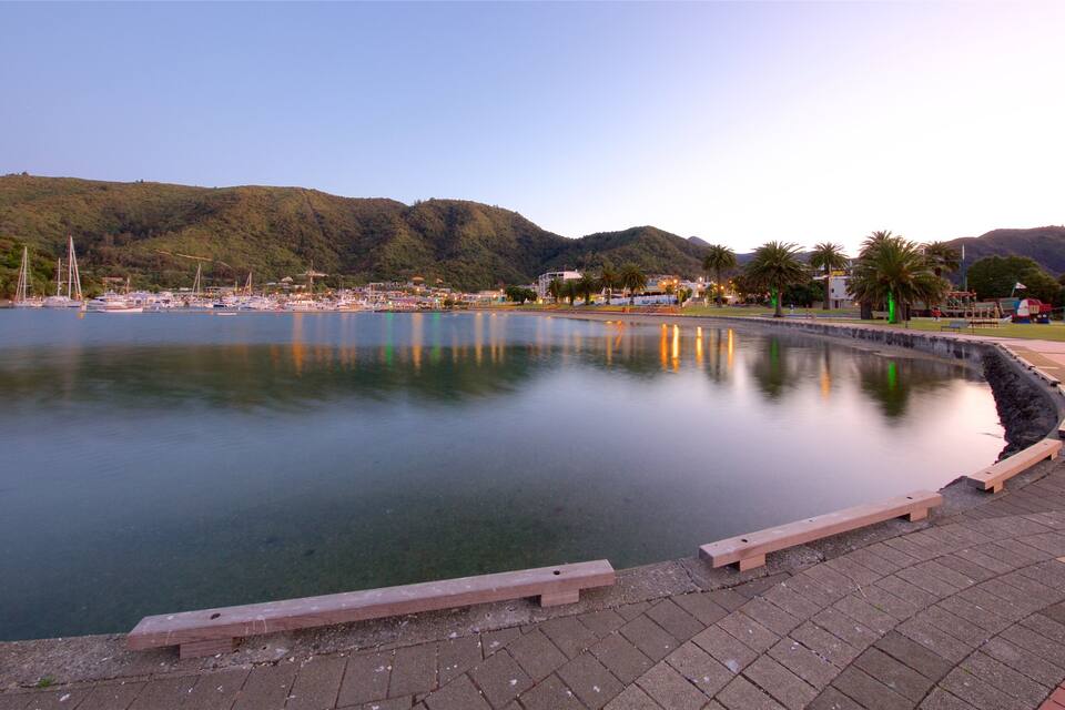 Un lago tranquilo con un banco de madera en primer plano, rodeado por un camino pavimentado. Al fondo, colinas y un puerto deportivo con barcos.