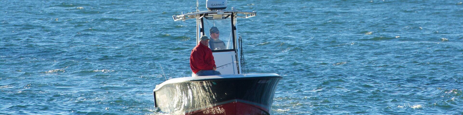 A small boat with a man in a red shirt on the water. There are multiple flags and a pier in the background.