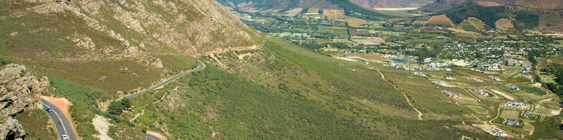 Una carretera sinuosa en la montaña, un valle con casas dispersas y una cadena de montañas al fondo.