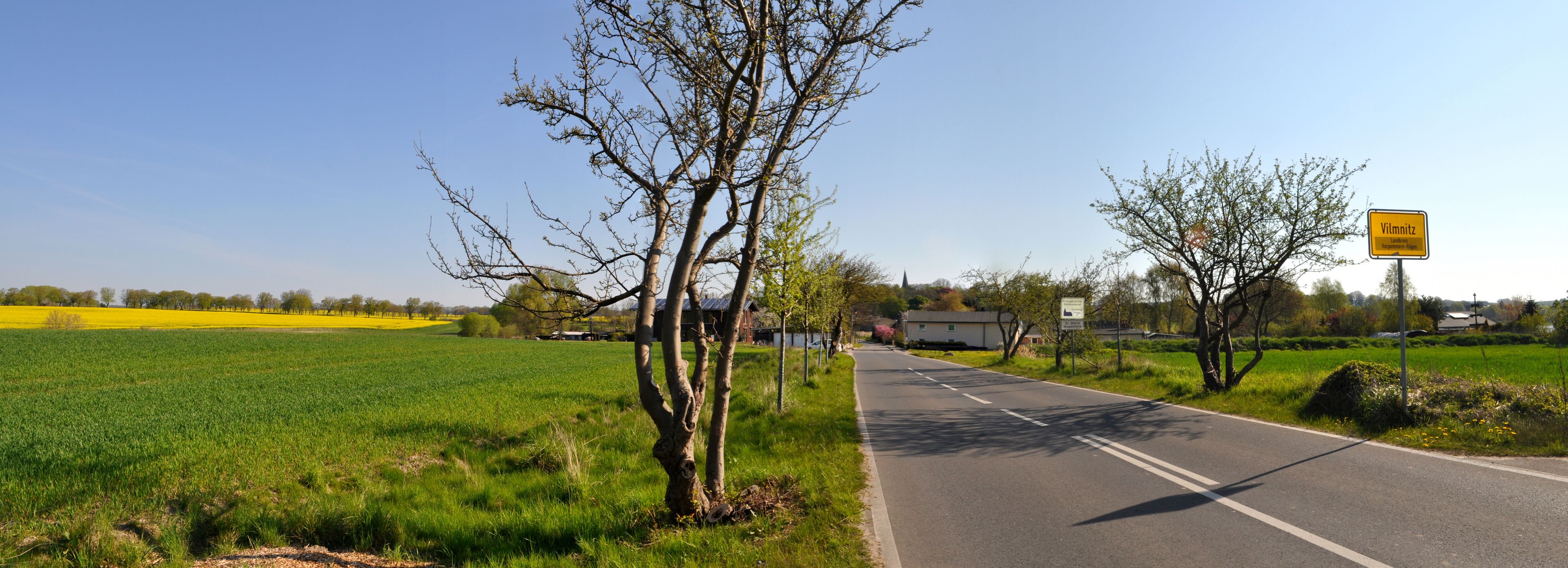 Eine ländliche Straße mit Feldern rechts und links, ein Schild für Vilmitz und ein Kirchturm in der Ferne.