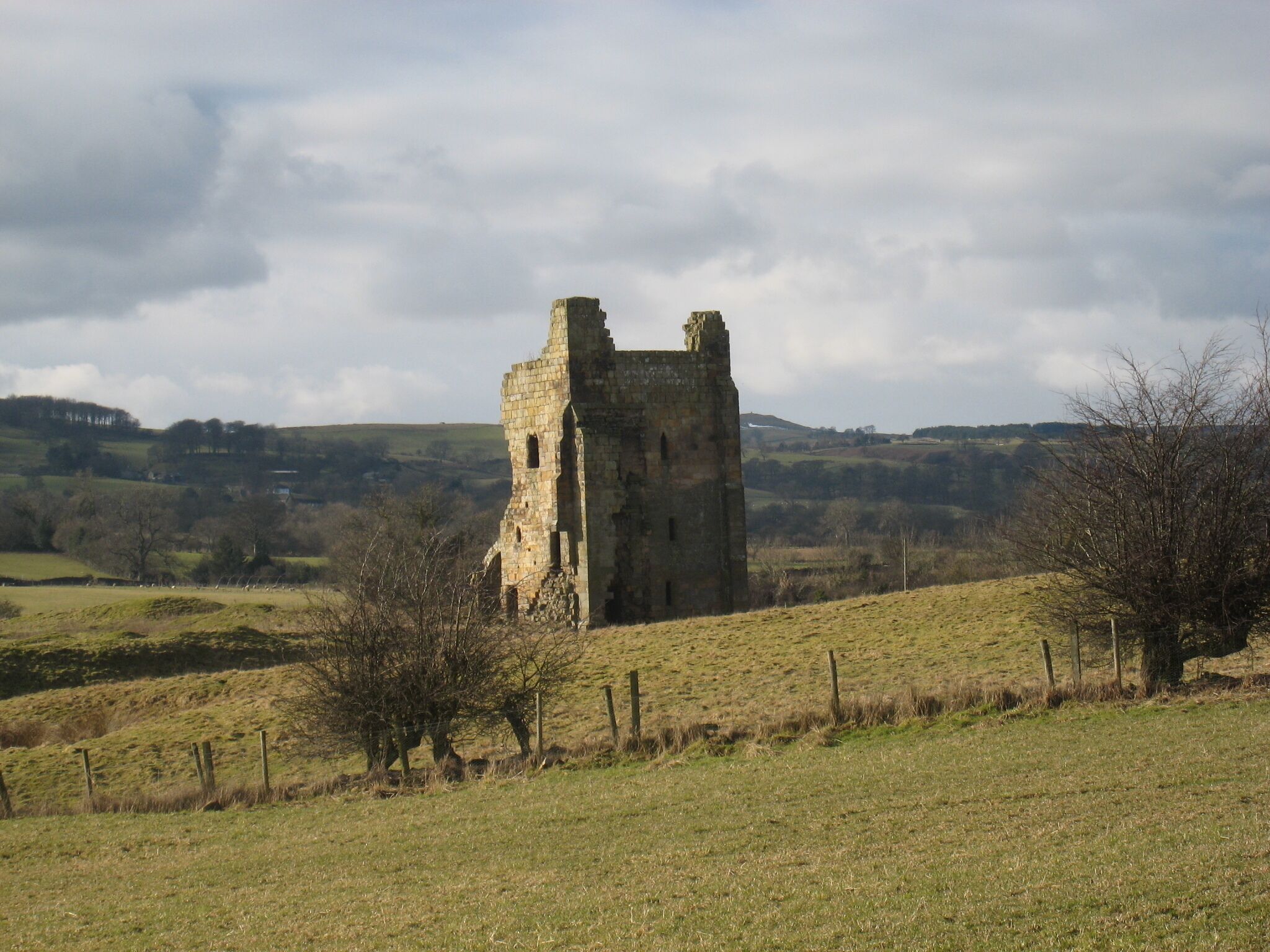 A medieval stone tower in a rural landscape with rolling hills and sparse trees.