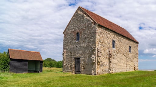 A stone building with a red roof, a small wooden structure, and a grassy field with trees in the background.