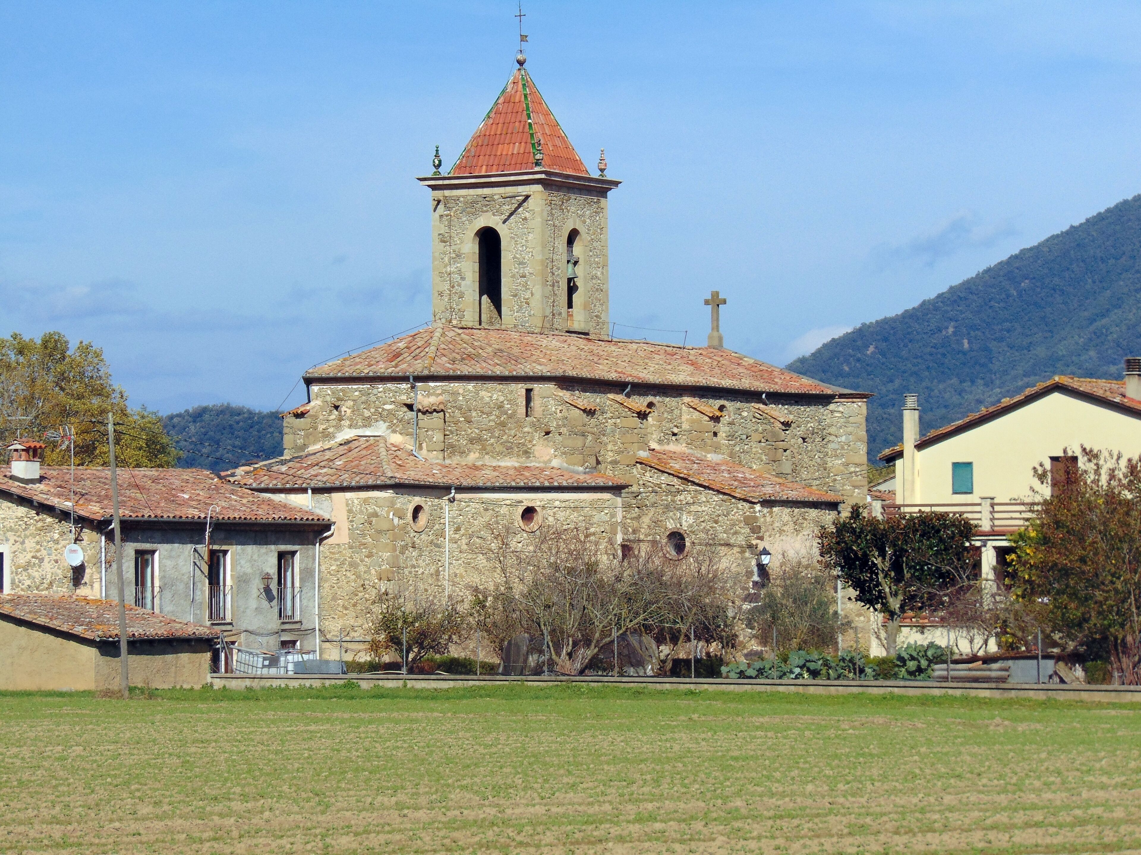 A stone church with a bell tower and a cross on top, surrounded by houses and a field.