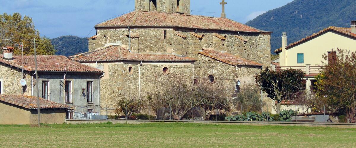 A stone church with a bell tower and a cross on top, surrounded by houses and a field.