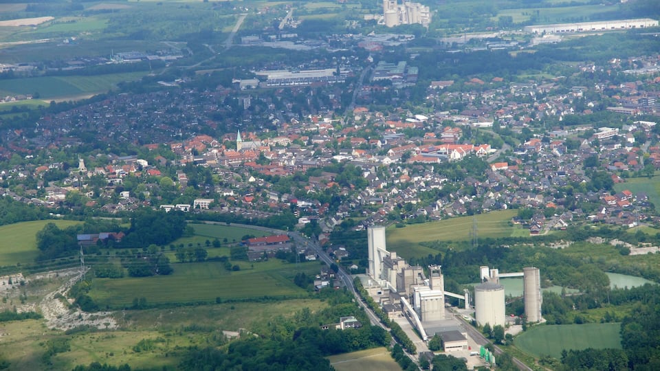 An aerial view of a town with a prominent castle, a large industrial complex, and surrounding green fields.