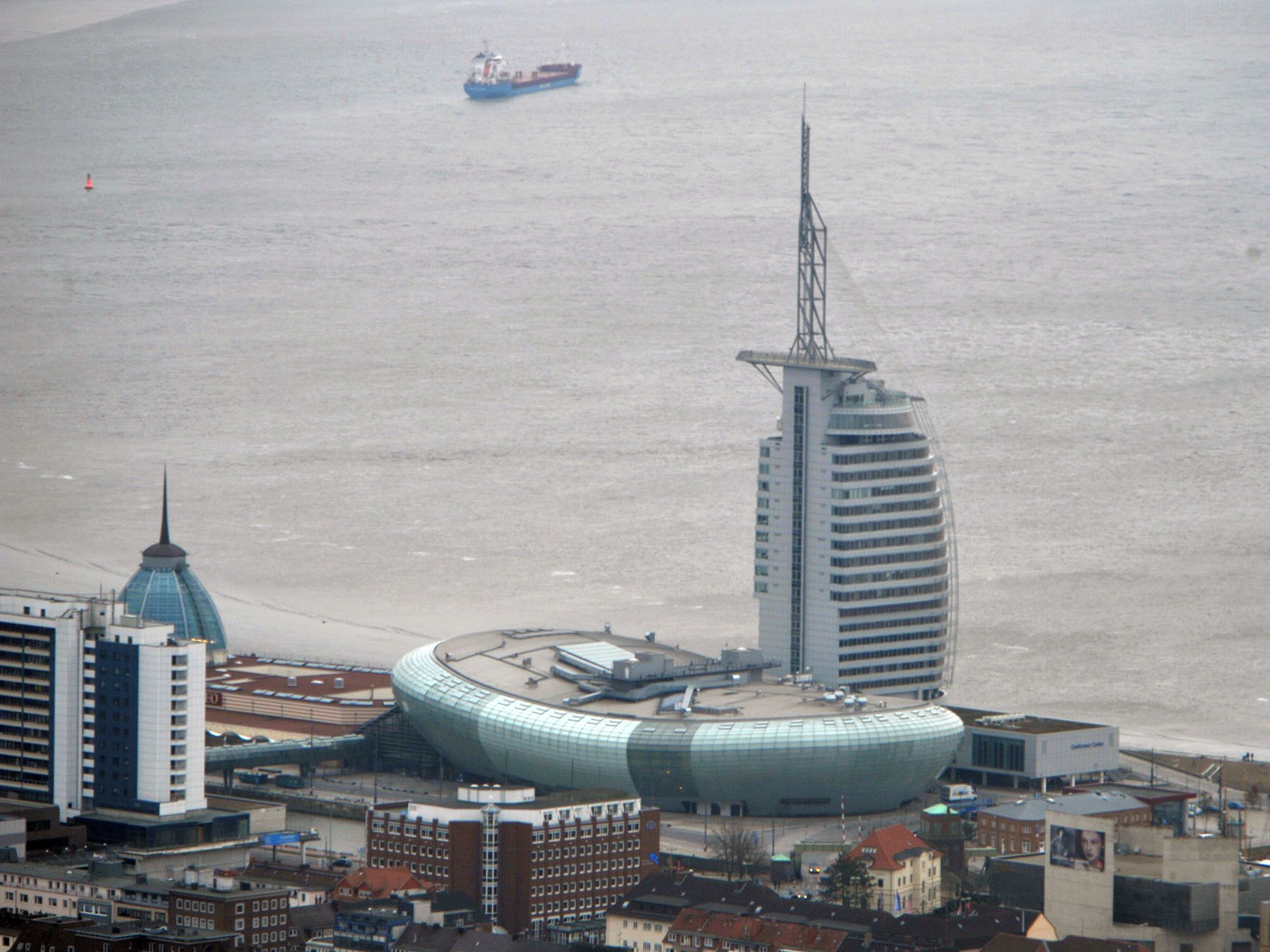 A coastal cityscape with a prominent modern building, a traditional tower, and a ship in the water.