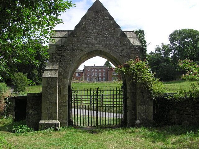 A stone archway with a metal gate, surrounded by greenery and a building in the background.