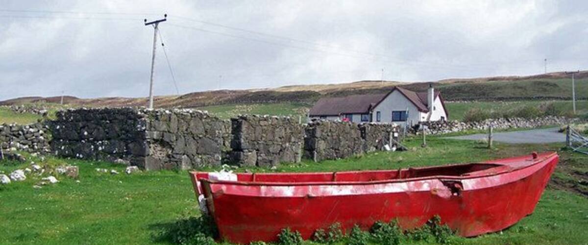 A red boat in a grassy field, a stone wall, a white house, and a hill in the background.