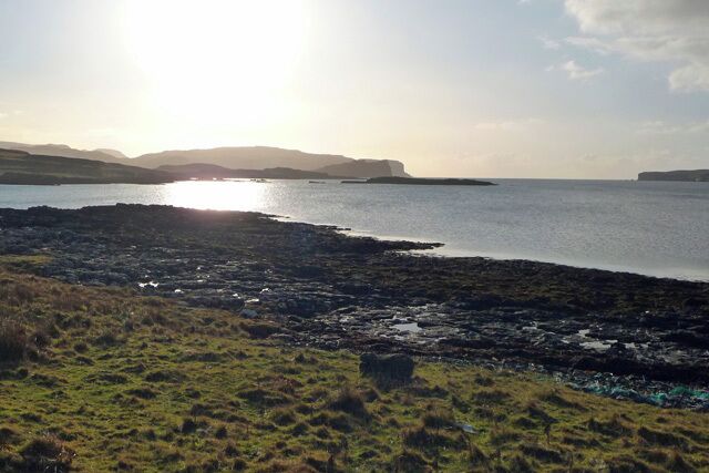 A coastal landscape with rocky shores, grassy areas, and distant hills under a clear sky.