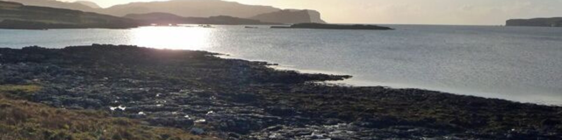 A coastal landscape with rocky shores, grassy areas, and distant hills under a clear sky.