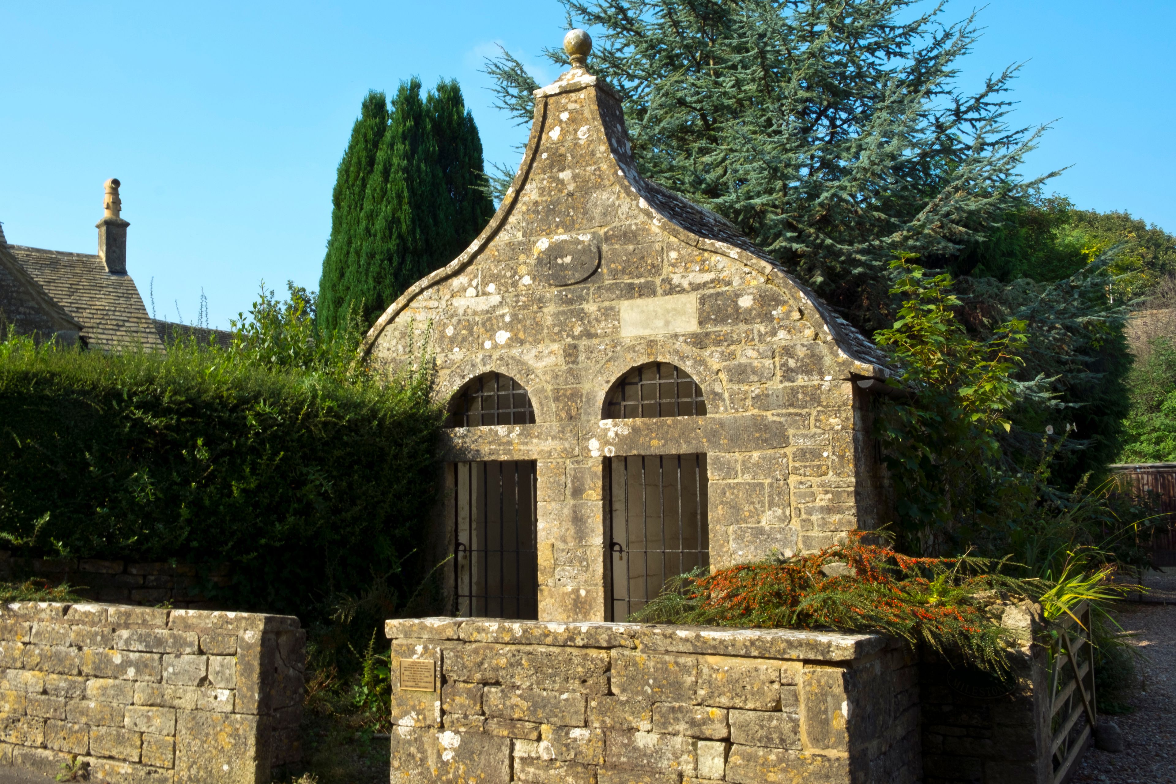 A stone building with a pointed roof, two arched windows, and a plaque.
