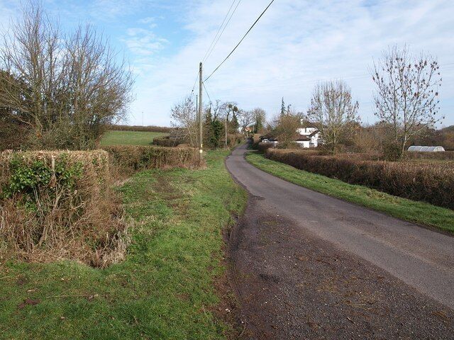 A narrow rural road curving through a countryside with hedgerows and a few houses in the distance.
