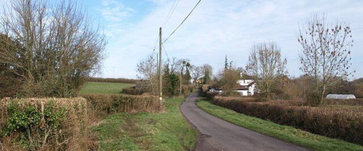 A narrow rural road curving through a countryside with hedgerows and a few houses in the distance.