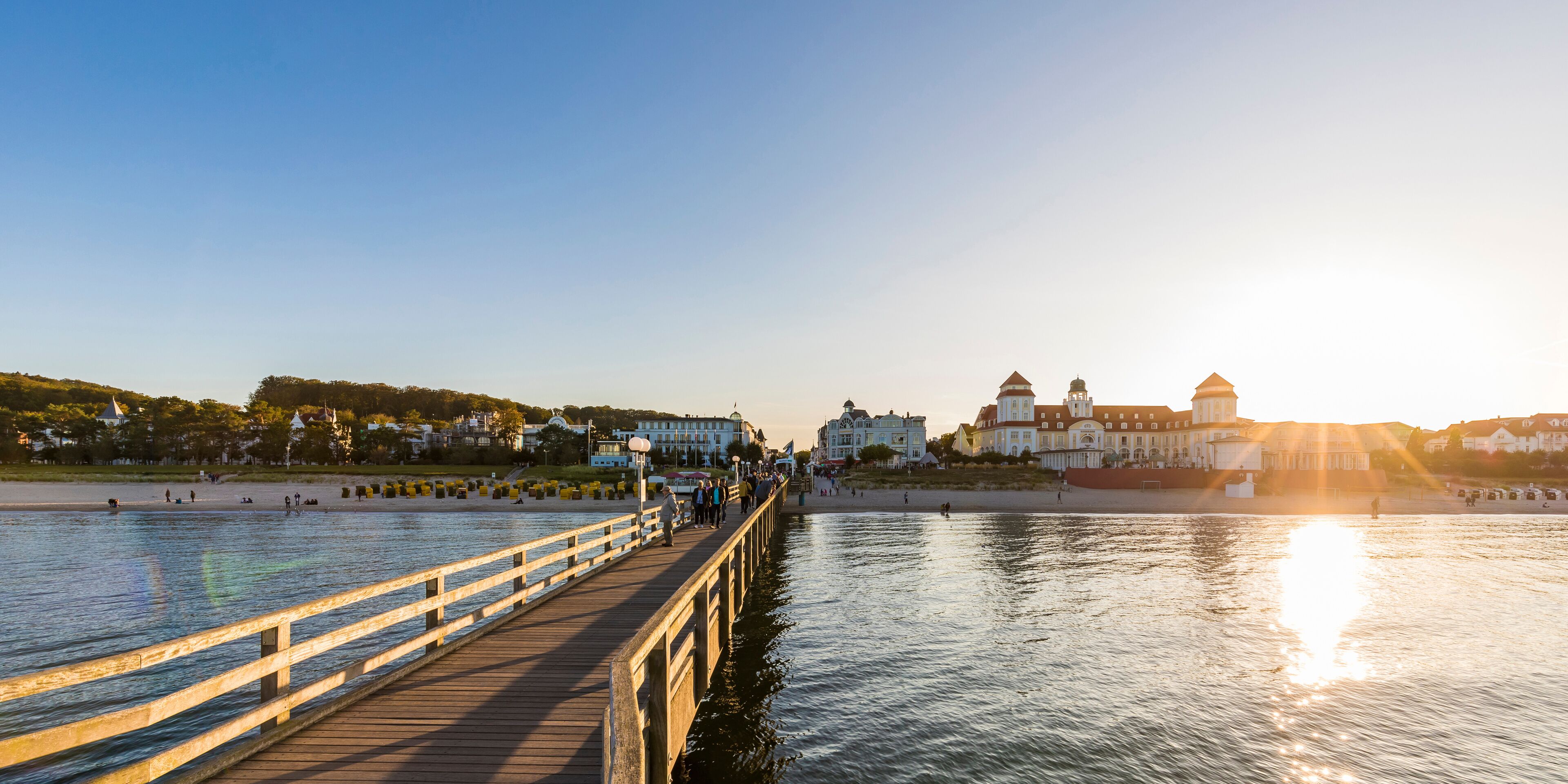 A wooden pier extending over calm water, with a sunset in the background and buildings on the opposite shore.