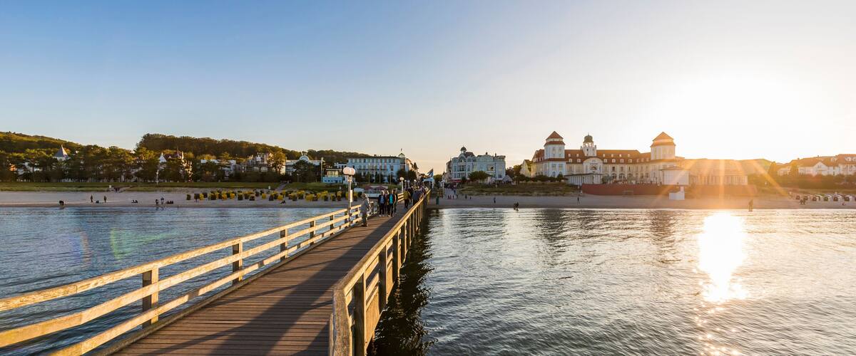 A wooden pier extending over calm water, with a sunset in the background and buildings on the opposite shore.