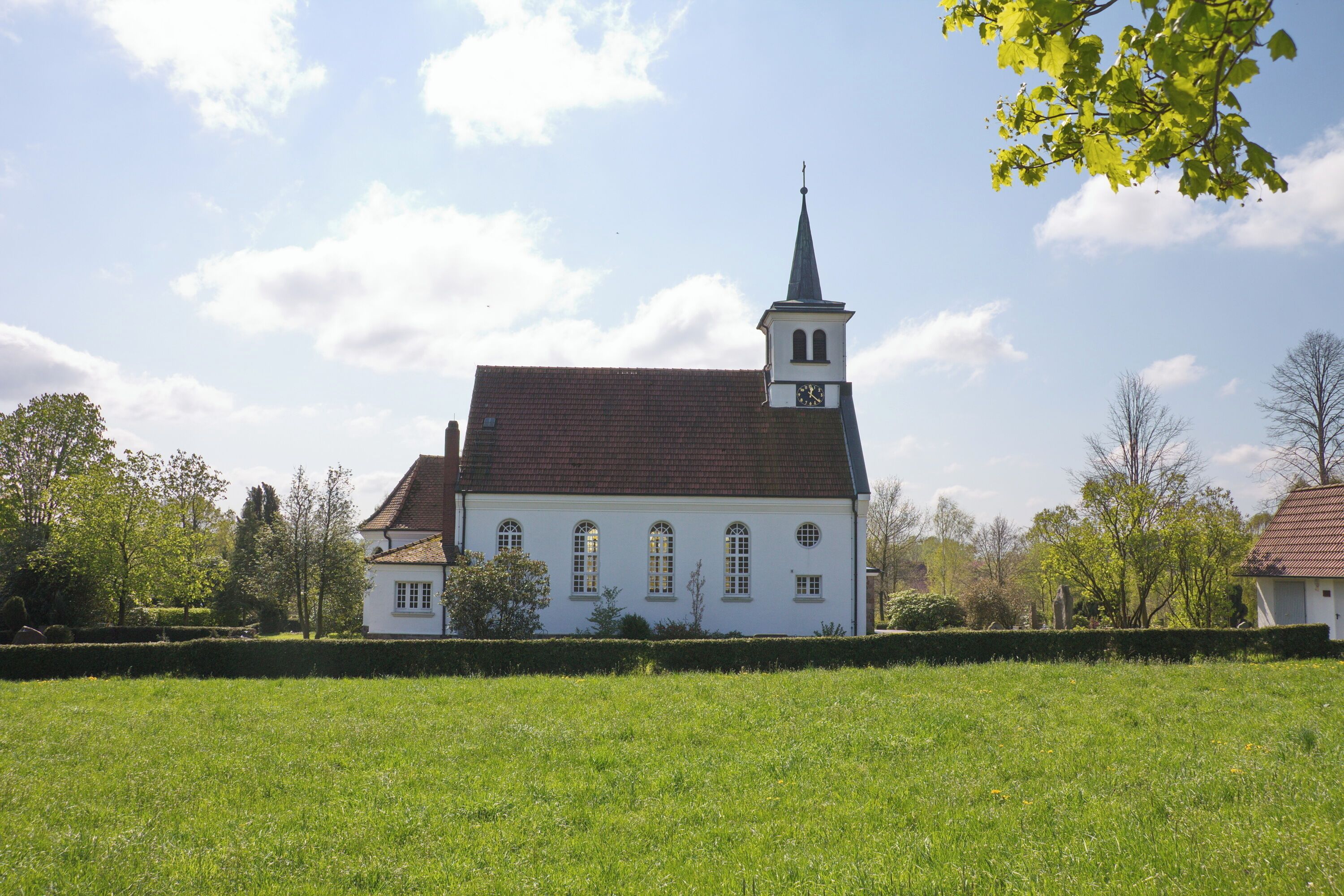 A white church with a steeple and a clock tower, surrounded by a green field and trees.